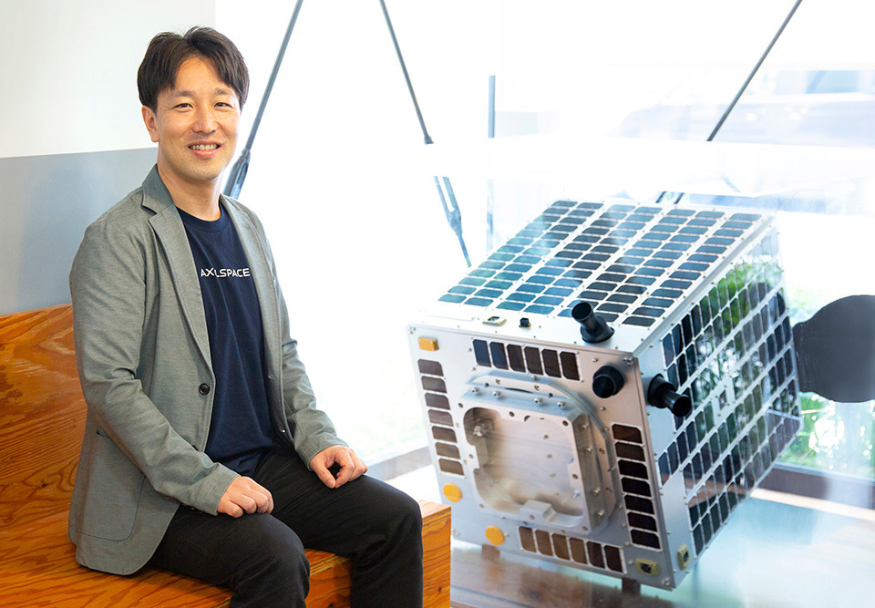 NAKAMURA Yuya, CEO of Axelspace, is sitting by a full-scale model of GRUS microsatellites.