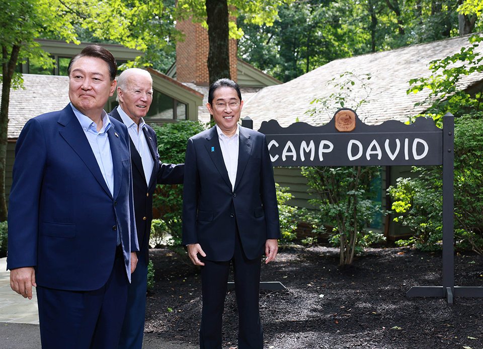 Prime Minister Kishida, Joe Biden, president of the United States, and President Yoon Suk Yeol, president of the Republic of Korea, standing beside the Camp David sign with lush greenery in the background.