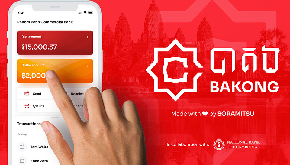 A photo of a hand operating a smartphone with the Bakong app open on the screen, red with a white logo and text in Khmer in the background.