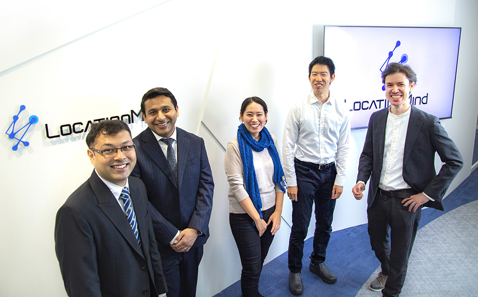 LocationMind team who are developing the PIVE (Poverty Impact and Vulnerability Evaluation) tool, including SHIBASAKI Mariko and and MIYAZAWA Satoshi, standing in front of a LocationMind sign on white wall.
