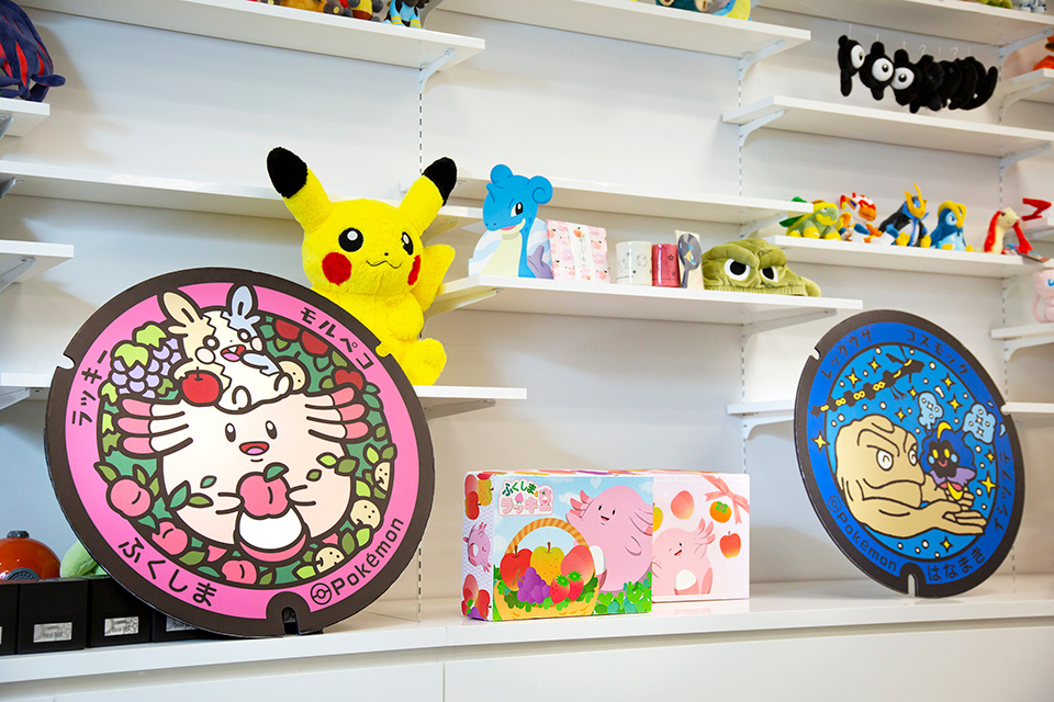 Two colorful Pokémon-themed maitenance covers, known as ‘Poké Lids,’ on a shelf with other Pokémon merchandise. 