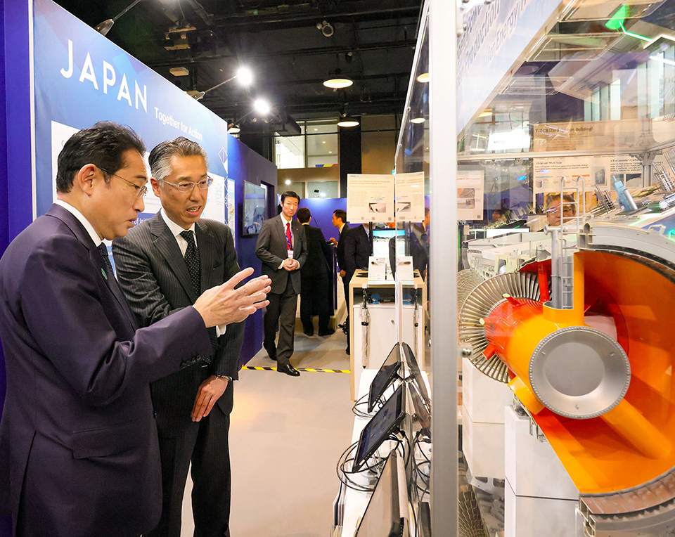 PM Kishida visiting a corporate booth at the Japan Pavilion at COP28 in Dubai, United Arab Emirates. A large, colorful mechanical exhibit visible at the right.