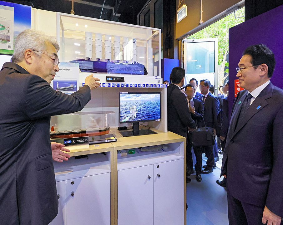 Prime Minister Kishida observing a display case containing models of technological equipment at the Japan Pavilion at COP28 in Dubai, United Arab Emirates. Another man is pointing towards the equipment alongside him.