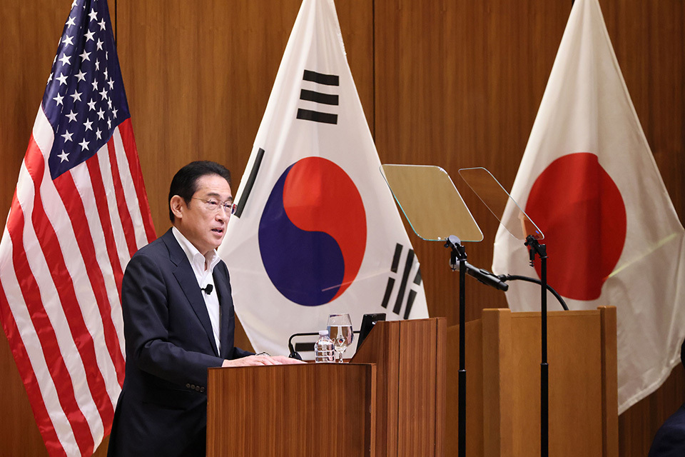 Prime Minister Kishida stands at a wooden podium delivering a speech, the flags of the United States, South Korea, and Japan in the background.