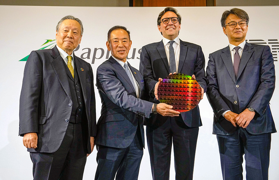 Four exectives of Rapidus and IBM, with two of them in the center jointly holding a large, colorful wafer.