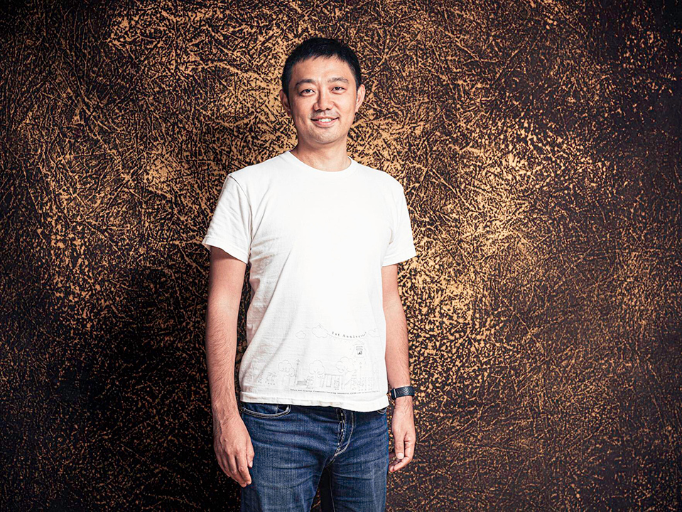 Sadoshima Yohei wearing a white t-shirt and dark jeans stands against a textured, golden-brown background.