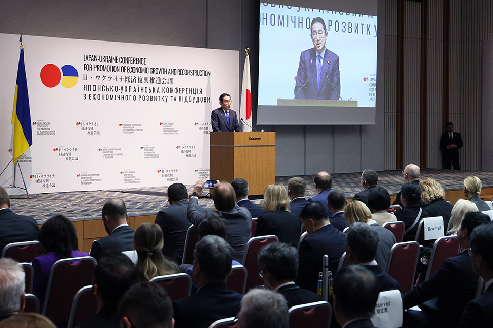 PM Kishida at the podium, attendees listen attentively, flags of Japan and Ukraine are in the background.