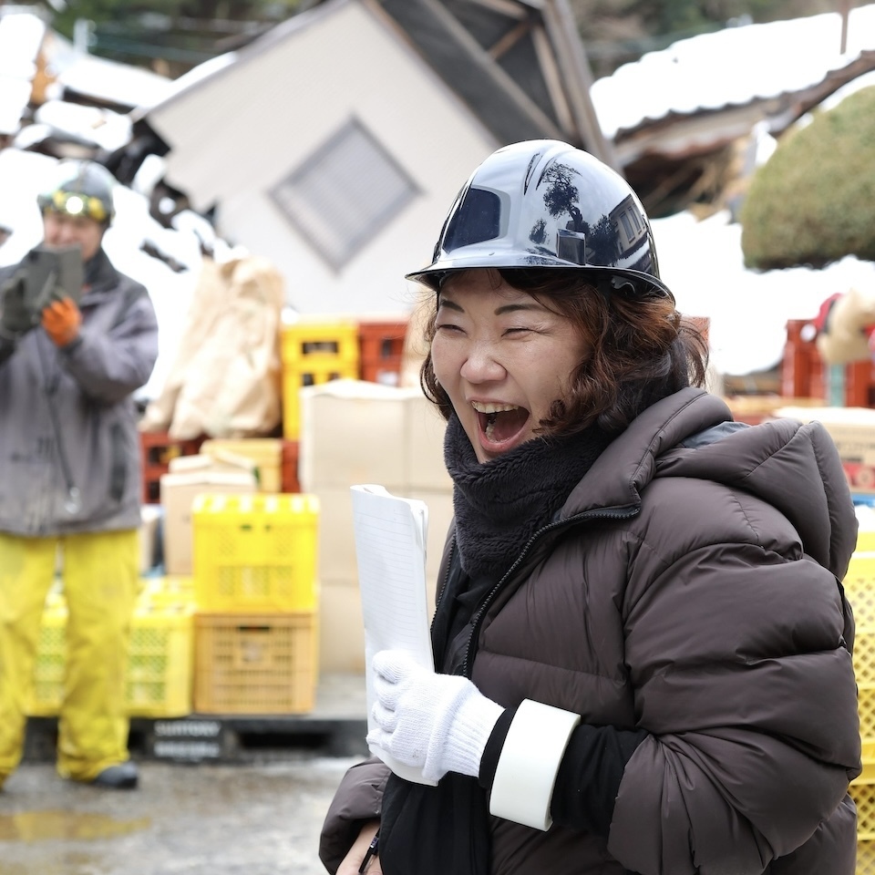 Joyful Kinshichi Seiko amidst debris, surrounded by stacked sake cases and rice bags in a disaster-stricken area.