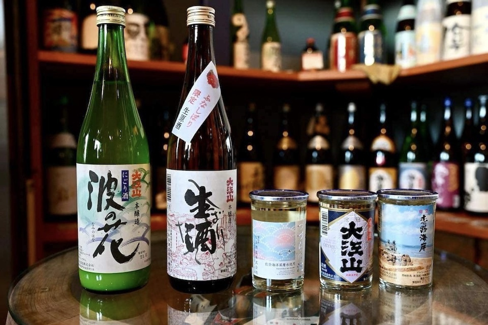 Two bottles and three glasses of sake are on a table, a shelf filled with sake bottles in the background.