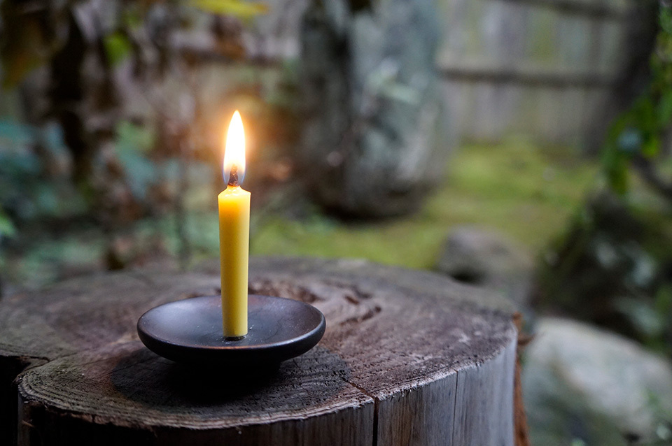 A lit candle in a black holder placed atop a weathered tree stump, illuminating a serene Japanese garden.
