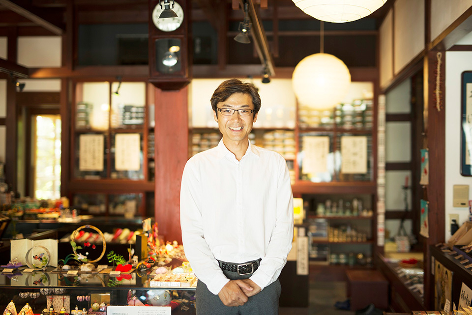 Takazawa in a white shirt and black pants standing in a well-lit, elegant Takazawa candle shop with many candles displayed.