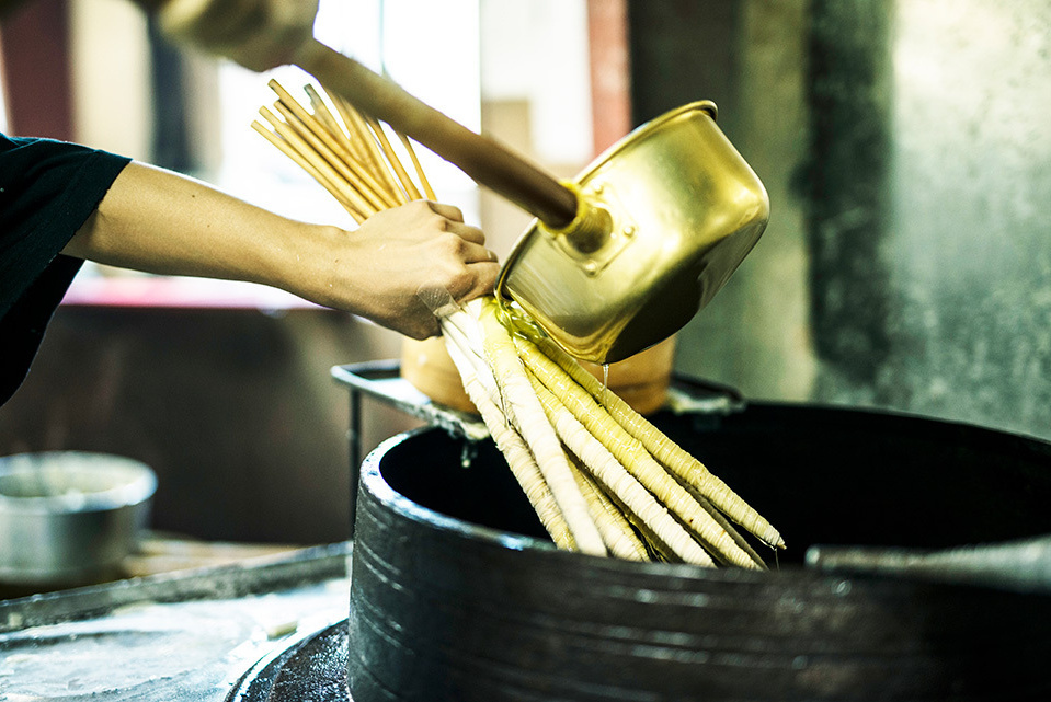 A person holding multiple long, uncoated wicks over a large vat, poring additional wax over them with a golden ladle.