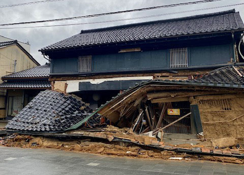 A damaged traditional Japanese house with its front wall and roof partially collapsed, exposing the wooden structure.