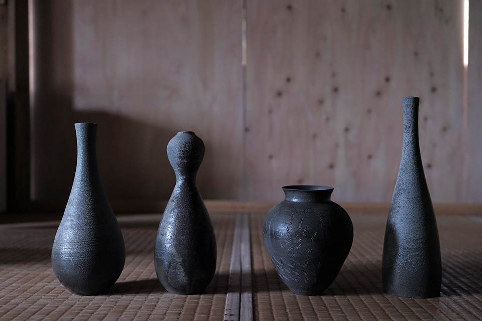Four unique pottery vases that are dark and have a rough texture, placed on a tatami mat.