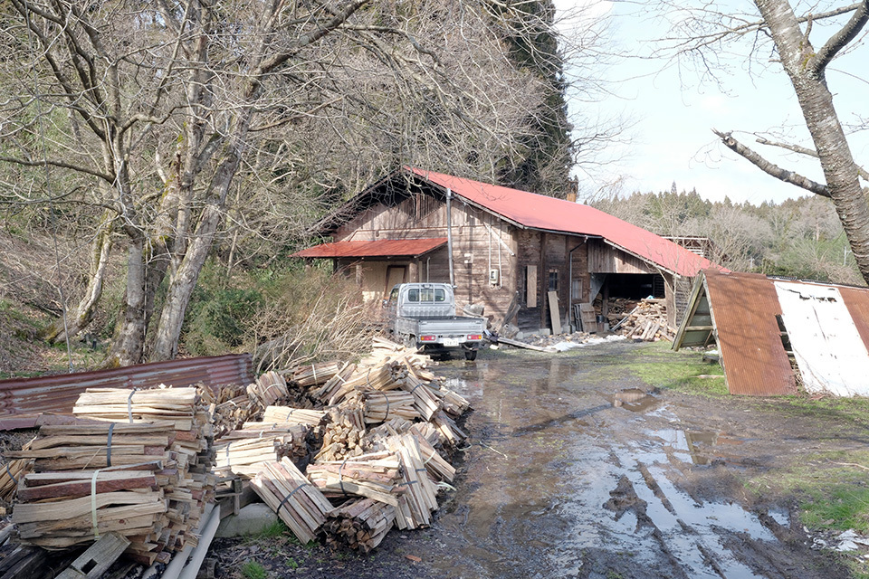 A rustic wooden building with a red roof, surrounded by piles of chopped wood and a parked vehicle.