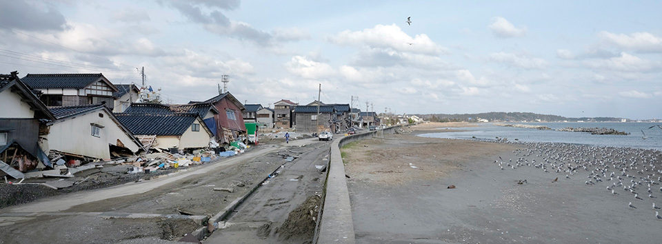 A coastal area showing signs of devastation with damaged houses and debris on one side, and a beach on the other.