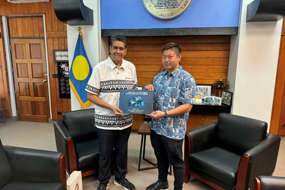 Two men posing in an office in front of the national flag of Palau, holding a plaque