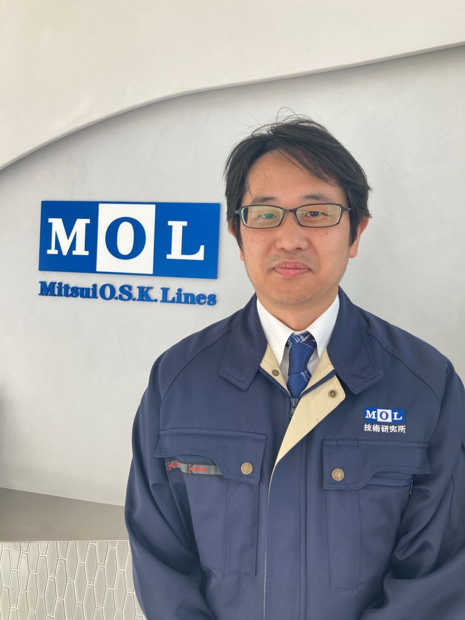 A Japanese man in a blue jacket and tie posing in front of the Mitsui O.S.K. Lines logo