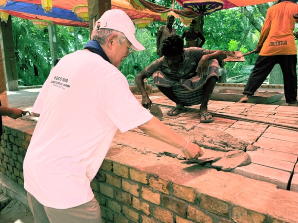 An older Japanese man troweling over non-fired bricks, watched by a worker in Bangladesh
