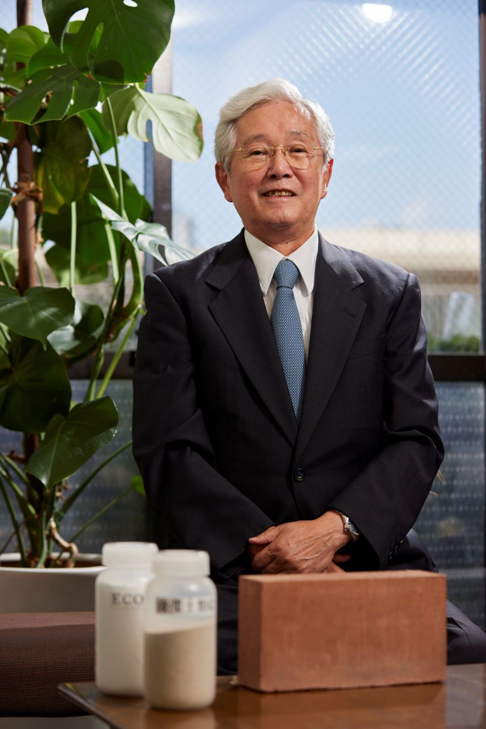 A white-haired Japanese man in a dark suit, standing behind a large brick and bottles of white powder