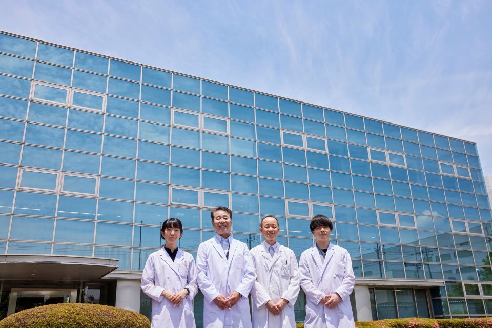 A team of four scientists in white lab coats posing in front of a modern, glass-fronted building