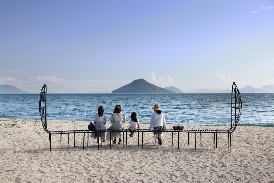 Three women and one child seated on a bench-like artwork, Place for Sea Dreamers, on a sandy beach overlooking the ocean and a island in the distance.