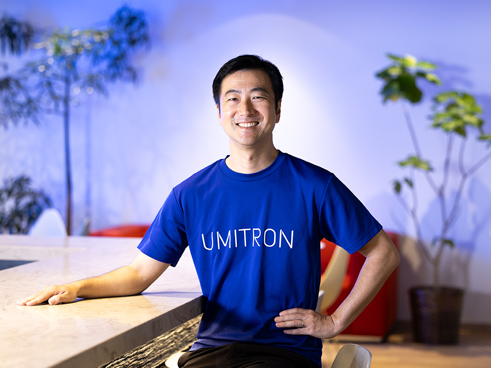A Japanese man in a blue shirt seated at a table in front of a lavender background