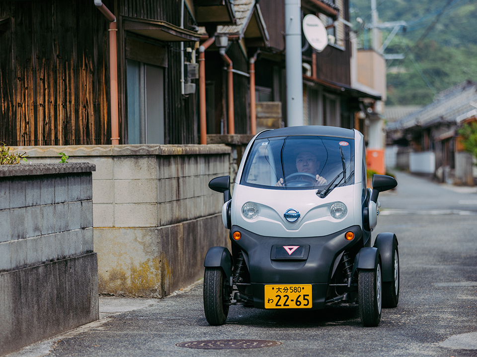 A small EV driving along a narrow road in a Japanese village